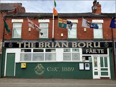A contemporary picture of the Brian Borou Irish Club. A two storey red brick building, decorated with flags. Four windows on the second storey, a large sign showing the name ‘Brian Borou’ across the fascia and a paining of an Irish Harp with the words. Es