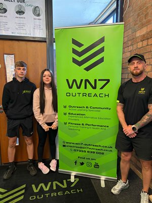 A young boy and girl stand next to each other, to their right is a large green sign that reads 'WN7 Outreach' and an older man is standing on the other side of the sign