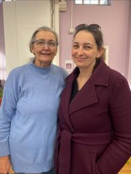Rachel and her mum stood in front of a light purple wall. Rachel is wearing a dark burgundy coat with sunglasses resting on her head, her mum is wearing a light blue long-sleeved top. A doorway and some wall fixtures are visible in the background.