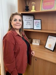 A lady stands beside a wooden shelving unit, wearing a red button-up jacket over a dark top. The shelves display framed prints with handwritten-style quotes, a small decorative light, two candles in transparent holders, and a few additional decorative ite